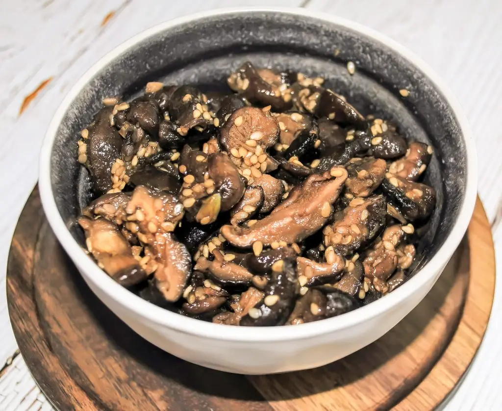 A white bowl of marinated mushrooms sitting on a round wooden platter.